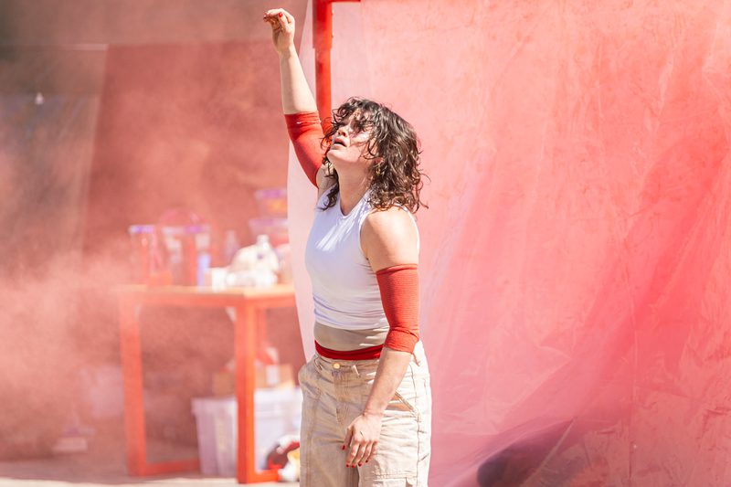 A white woman with black curly hair is stood in front of a red pole. She is surrounded by red smoke. Her arm is stretched above her head and she looks distressed.