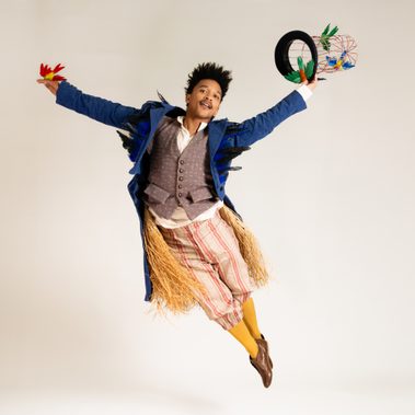 A performer jumps with both arms raised, wearing a blue tailcoat and yellow trousers and socks