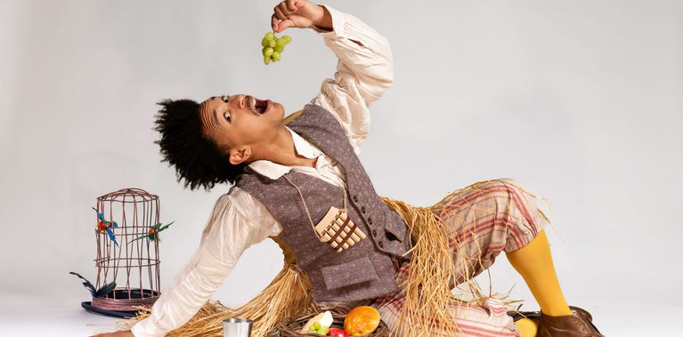 A performer dressed in a waistcoat and shirt and yellow trousers and socks, dangles some grapes above their mouth