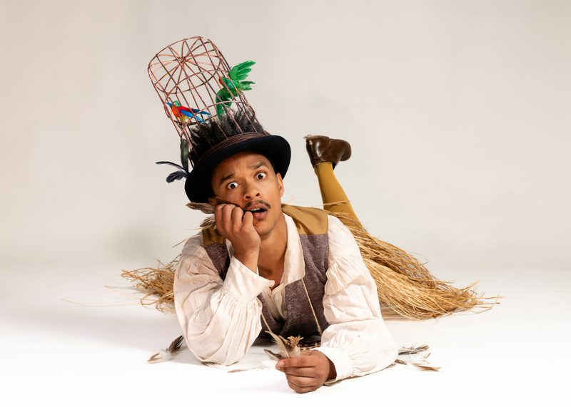 A person lies on the floor wearing a tall birdcage hat with artificial birds, holding a feather and looking surprised.
