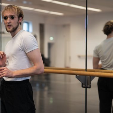 A person wearing a white t-shirt and small round sunglasses stands in a dance studio, positioned by a wooden ballet barre in front of a large mirror that reflects their back.