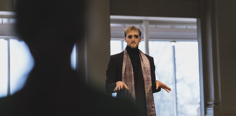 A person wearing round sunglasses, a dark suit, and a patterned scarf stands indoors with hands raised mid-gesture, framed by columns and large windows, with a blurred figure in the foreground.