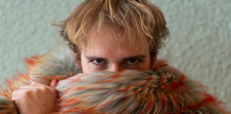 Close-up of a person with short blond hair staring intensely at the camera while partially covering their face with a large, multicolored fur garment streaked with orange and gray.