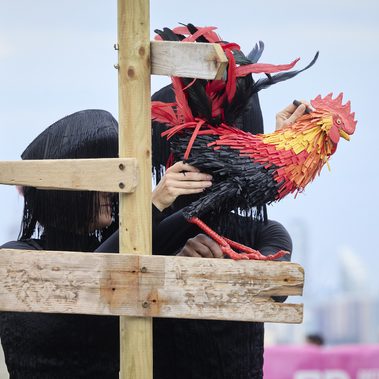 Two performers dressed in black manipulate a rooster puppet beside a weathered wooden signpost.