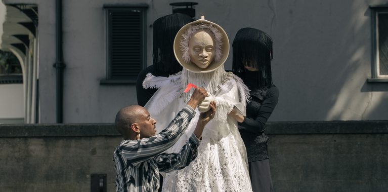 A performer reaches toward a masked figure in an elaborate white costume, supported by two puppeteers dressed in black standing behind it.