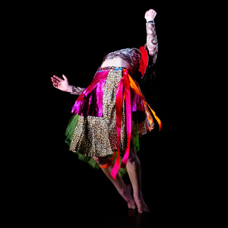 Person wearing a skirt with ribbons in pinks and oranges and leopard print hanging off. They are leaning backwards so their head is not visible. The background is black.