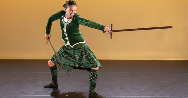 Person on stage wearing a green traditional Scottish outfit holding a sword in both hands and pointing one out.