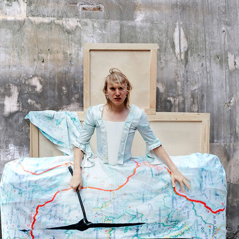 Anders Duckworth stands in a costume that resembles a 15th century dress with a greatly exaggerated skirt made out of a map.
