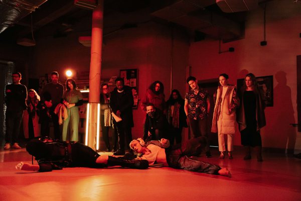 The room is lit with red lights. Audience members are standing watching two performers on the floor. One is wearing headphones and sports clothes, and is holding the leg of the second performer who is wearing platform boots and an all black outfit and their faced covered