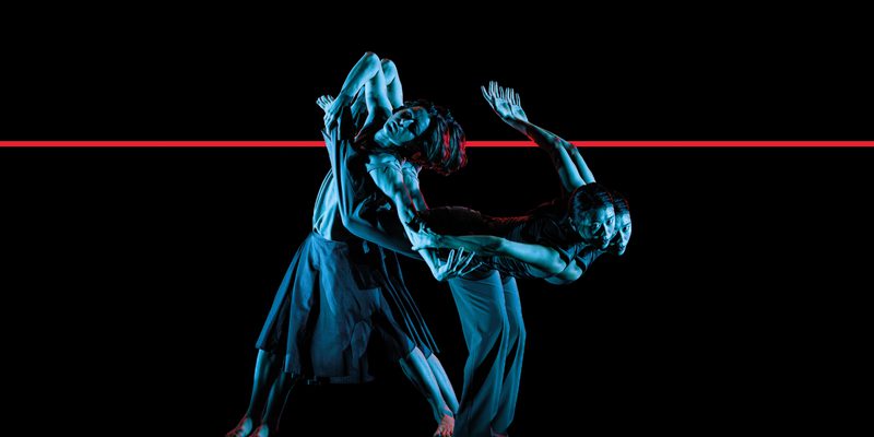 Two dancers on a black background lit with pale blue light wind together in front of a red strip light.