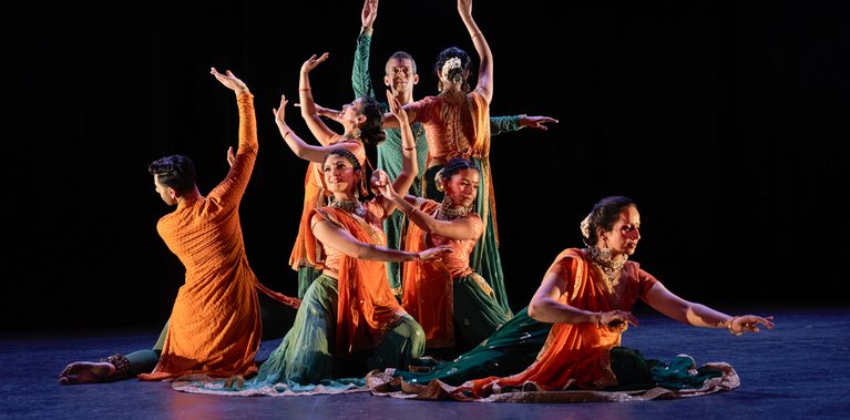 Seven Kathak dancers: two male, five female, in bright saffron orange and sea green costumes, trimmed with gold, forming a tableau. All the dancers are making slightly different gestures, some seated, some standing. The effect is of a complex organism. The women have gold jewellery and white flowers in their hair. The stage is hung with black and the light tints the floor blue