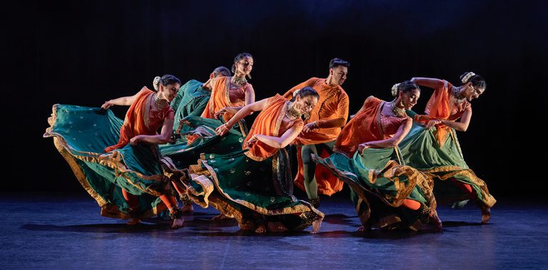 Seven Kathak dancers, five female two male, in bright saffron orange and sea green costumes, caught in the middle of a dramatic swirling step performed in unison. They are inclining towards their left and swirling their arms and skirts towards their right, with their left legs bent and turned inward. The women have gold jewellery and white flowers in their hair. The stage is hung with black and the light tints the floor blue.