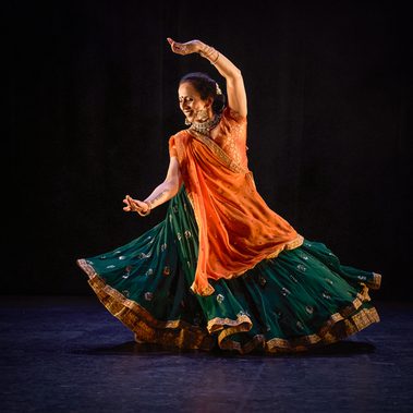 A female Kathak dancer makes a classical gesture as she comes out of a spin. Her left arm is raised and curved back over her head, her right is extended towards the downstage corner. Her hands are soft and she smiles towards her extended hand. She is wearing a dark green skirt with gold details that swirls around her, with an orange and gold top and pinned dupatta/scarf. She has gold Indian jewellery and white flowers in her hair. The stage is hung with black and the light tints the floor purple.