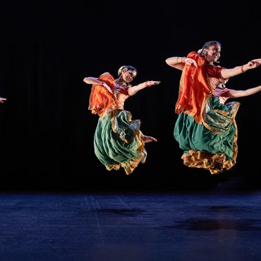 Seven Kathak dancers: two male, five female, in bright saffron orange and sea green costumes, caught mid-leap. They are grouped three towards the back, and four towards the front, but are all dancing in unison. Their legs are tucked up under them, and their arms are all extended in the same direction. Those whose faces are visible are smiling. The women have gold jewellery and white flowers in their hair. The stage is hung with black and the light tints the floor blue.