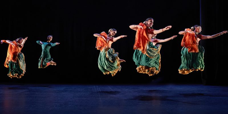 Seven Kathak dancers: two male, five female, in bright saffron orange and sea green costumes, caught mid-leap. They are grouped three towards the back, and four towards the front, but are all dancing in unison. Their legs are tucked up under them, and their arms are all extended in the same direction. Those whose faces are visible are smiling. The women have gold jewellery and white flowers in their hair. The stage is hung with black and the light tints the floor blue.