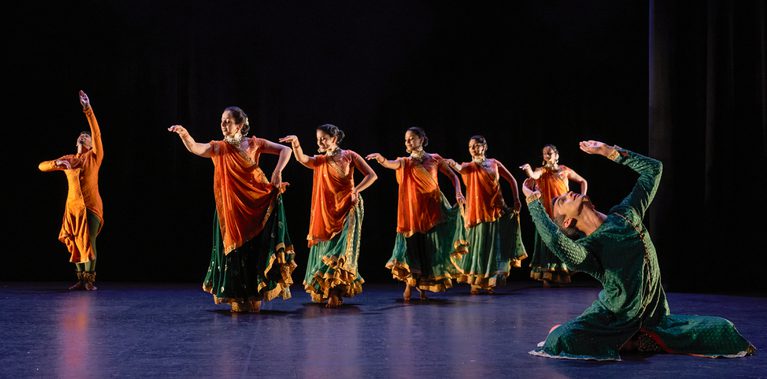 Seven Kathak dancers: two male, five female, in bright saffron orange and sea green costumes, performing. The five female dancers are processing diagonally across the stage, holding their skirts out with one hand and gesturing forwards with the other. In the back, a male dancer with orange stands, reaching upwards with one arm and his gaze. In the front a male dancer in green leans backwards from a seated position with both arms raised. The stage is hung with black and the light tints the floor blue.