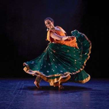 A female Kathak dancer swirls her skirts with her hands. She smiles towards her extended eg. She is wearing a dark green skirt with gold details, with an orange and gold top and pinned dupatta/scarf. She has gold Indian jewellery and white flowers in her hair. The stage is hung with black and the light tints the floor blue. Four female Kathak dancers gesture in unison from a seated tableau position with their right arms extended across their bodies. In the background, two male dancers make a mirrored pose, with opposite arms extended towards each other, and opposite legs extended away. All the dancers wear bright saffron orange and sea green costumes trimmed with gold. The women have gold jewellery and white flowers in their hair. The stage is hung with black and the light tints the floor blue.