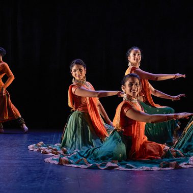 Four female Kathak dancers gesture in unison from a seated tableau position with their right arms extended across their bodies. In the background, two male dancers make a mirrored pose, with opposite arms extended towards each other, and opposite legs extended away. All the dancers wear bright saffron orange and sea green costumes trimmed with gold. The women have gold jewellery and white flowers in their hair. The stage is hung with black and the light tints the floor blue.