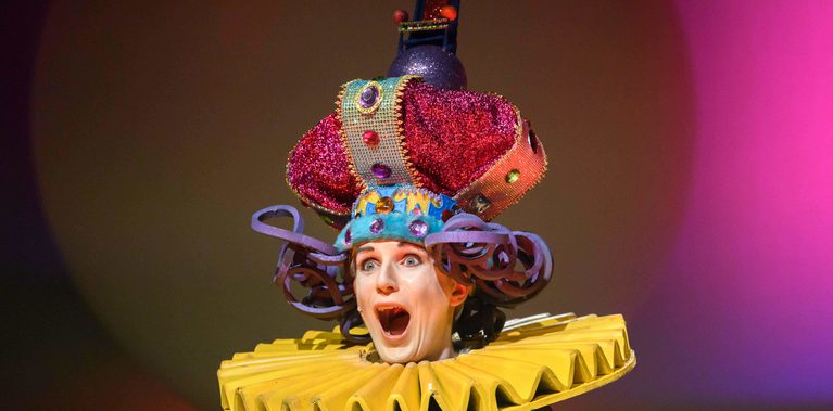 A performer wearing a large colourful crown and yellow collar is pointing to the audience. They are coming out of a large white ball. The lighting on the backdrop is bright pink.