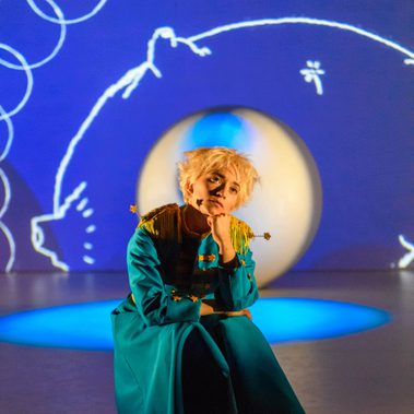 A performer dressed as The Little Prince sits on a stage, looking to the sky. Behind them is a large white ball and white graphic illustrations from the book projected on a blue backdrop.