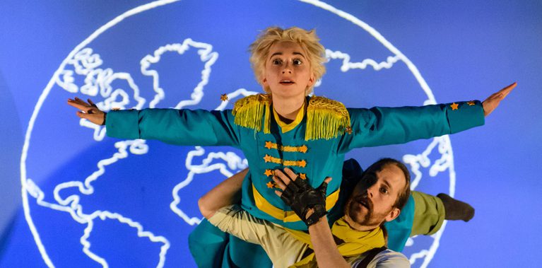 A performer dressed as a Pilot is holding another performer dressed as The Little Prince in a lift over there shoulder. The Little Prince has both arms stretch out wide and is looking up. There is a blue backdrop with a white graphic of the world behind them.
