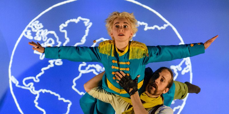 A performer dressed as a Pilot is holding another performer dressed as The Little Prince in a lift over there shoulder. The Little Prince has both arms stretch out wide and is looking up. There is a blue backdrop with a white graphic of the world behind them.