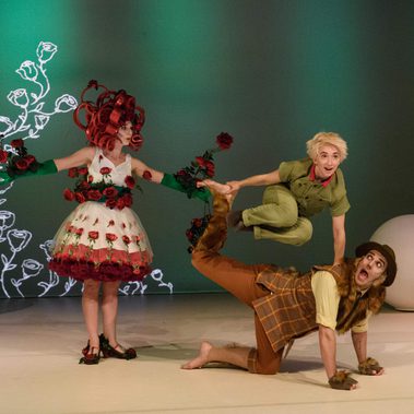 Three performers are on a white stage with a green backdrop. One is dressed as a rose and is pointing at The Little Prince who is wearing a green jumpsuit, jumping over a performer wearing a fox costume.