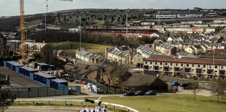 a dancer is tossed in the air by a group of people all looking up at her, arms stretched up to catch her. They are on a Derry street with the panoramic hillside streets of bogside behind them.