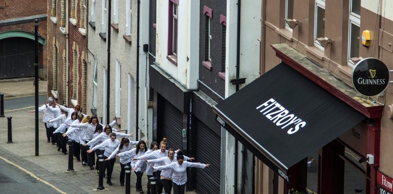 A line of white shirted dancers, arms outstretched as if walking a tightrope, walking up a city street in Derry, just passing a bar that sells Guinness.