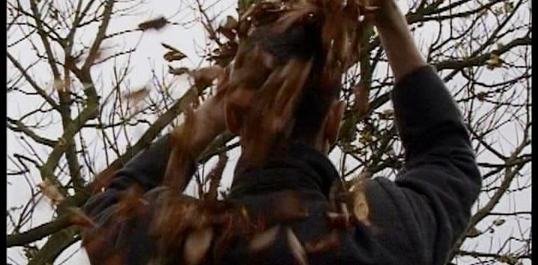 A man in a dark grey overcoat stands with his back to us in a wintery leafless wood, his hands are up over is head amongst the dry crisp autumnal leaves that are falling from his head and flying in the wind