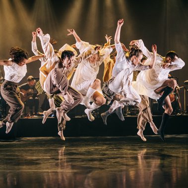 Dancers in white dresses gracefully leaping in the air, showcasing their elegance and skill. In the back ground there is a live band with dark warm lighting.