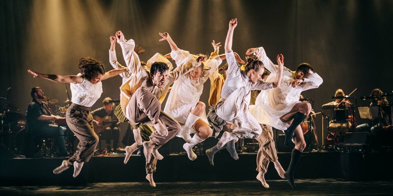 Dancers in white dresses gracefully leaping in the air, showcasing their elegance and skill. In the back ground there is a live band with dark warm lighting.