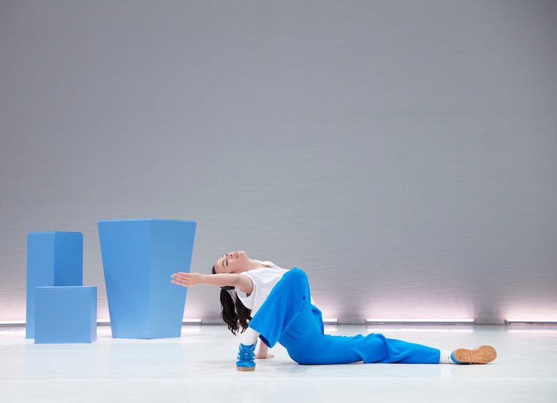 There is a white background and three blue cubes in the corner of the stage. A dancer is in the centre of the stage wearing a wearing top and blue trousers and trainers.
