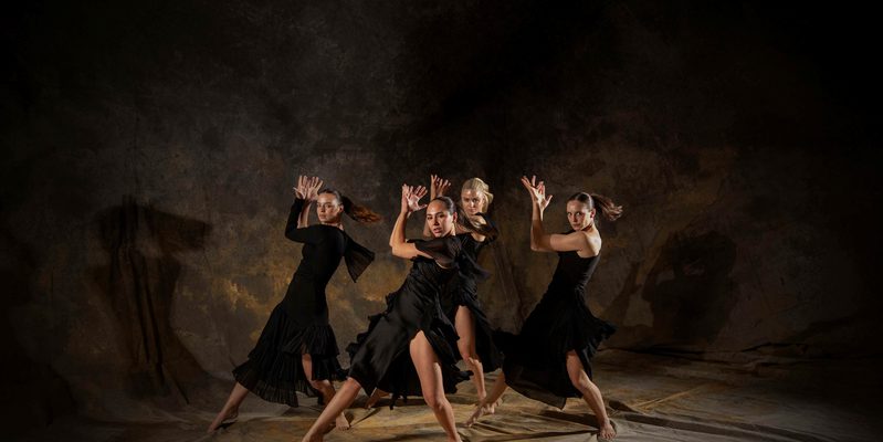 Four dancers in the same dynamic twisted position, hands in an expressive position near their faces. They are against a mottled fabric background.
