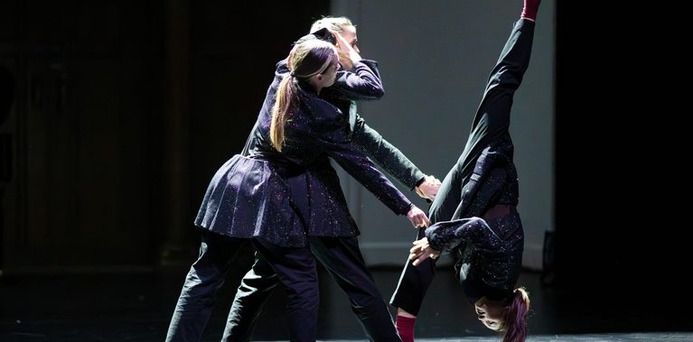 Two dancers hold another while they are doing standing splits at an unstable angle. They are wearing black sparkly outfits and maroon coloured socks.