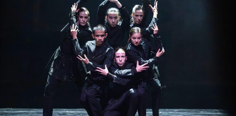 Six dancers in sparkly black outfits stand together making a circle with their arms.