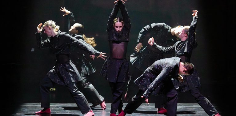 Five dancers are in different poses around a central figure who facing the camera and holding a straight posture with their hands crossed above their head. One dancer faces the central figure while the others face away.
