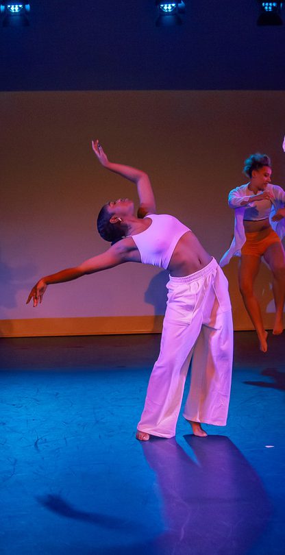 A dancer at the centre of the stage leans back, her arms held gracefully in the air above her head. Around her five dancers leap into the air in a circle.