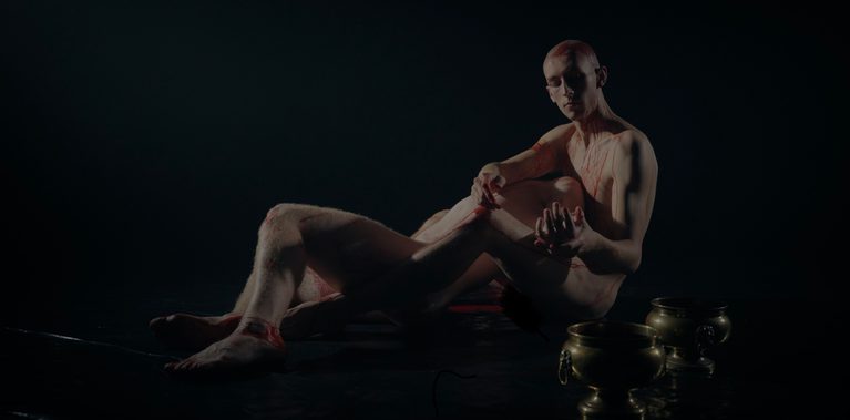 Two nearly identical looking naked men lie together on a black dance floor. Two golden vessels are on the floor in the foreground.