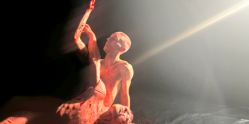 Two nearly identical looking naked men lie together on a black dance floor covered in blood. One lies in the other's lap while the other props themselves in a seated position gazing up at their outstretched hand.