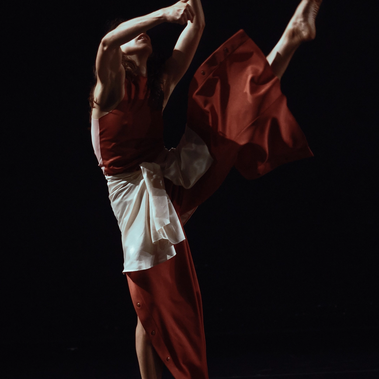 A person performs a dynamic contemporary dance on a dark stage. They are captured in mid-motion, balanced on one foot with the other leg extended high to the side. They wear a long, rust-red garment with a white fabric wrap around the waist. Their arms are raised toward the upper left, with one hand open and reaching, while their head is tilted back. Dramatic lighting highlights the dancer's form and the flowing textures of the fabric against the solid black background.