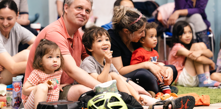 A close-up view of families sitting on a green rug. The children are focused forward with expressions of wonder and curiosity.