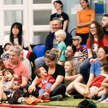 A group of adults and children sit closely together on a green rug and chairs in a brightly lit room. They are all looking toward the left with expressions of engagement and joy, suggesting they are watching a performance. Several parents are holding toddlers, and the atmosphere is warm and communal.