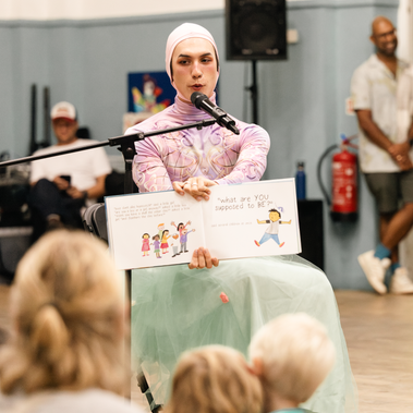 A performer wearing a pink hooded top and a light green tutu sits behind a microphone to lead a storytelling session. They are holding open a children's picture book to show the illustrations to the seated crowd. The performer is captured mid-sentence as they read aloud, while other adults in the background watch the performance.