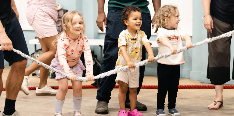 Three young children stand in a row, smiling as they hold onto a long, thick white rope. They appear to be participating in a group activity or game indoors, with adults standing nearby in a relaxed setting.