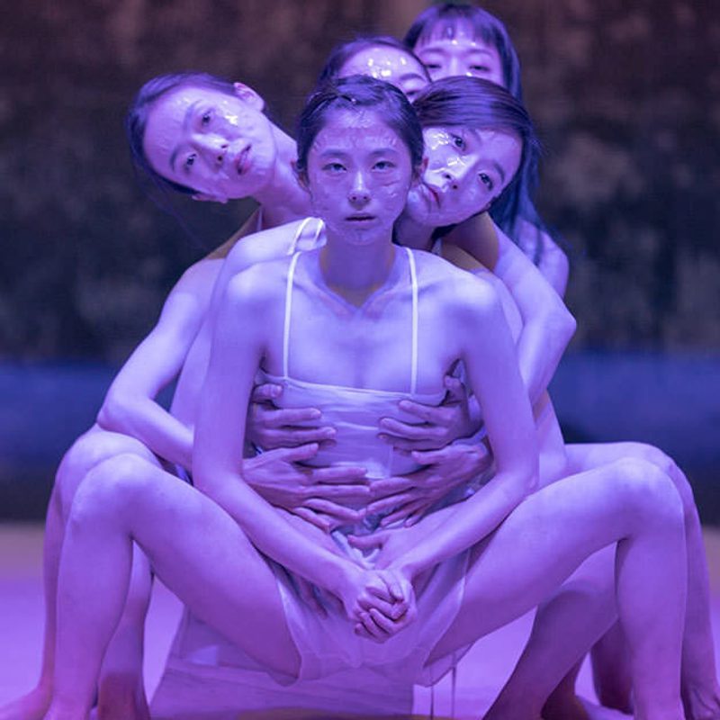 5 performers crouching one behind another. They have pink lighting on them and a clear sheath that is wrinkled over their faces..