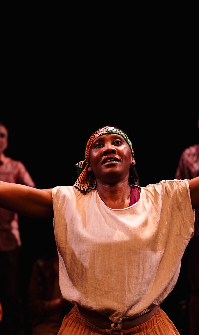 In the background, a group of performers are on stage. A female artist is holding out her arms wide, looking up.
