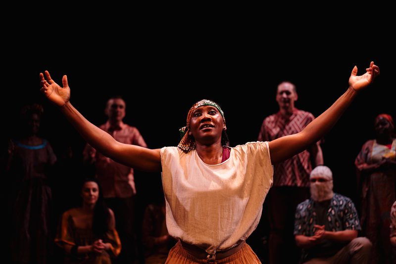 In the background, a group of performers are on stage. A female artist is holding out her arms wide, looking up.