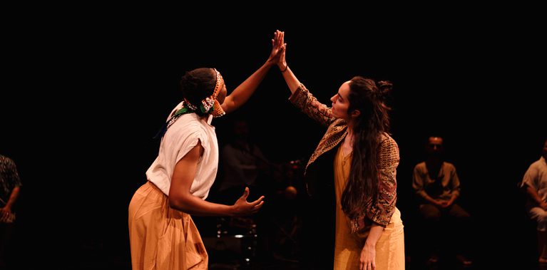 Two female performers are facing each other. They are holding each others hand and reaching up.