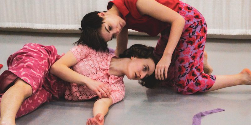 The image features two white female dancers with short dark hair. The floor and the background are both white. They are both wearing pink patterned trousers and short-sleeved blouses. One of the dancers is lying on the floor, while the other dancer is kneeling and bending over them with their head gently touching the other dancer’s shoulder.